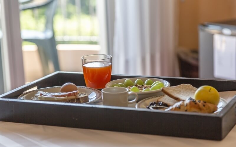 Breakfast tray with orange juice, coffee, grapes, boiled egg, bacon, toast, pastry, and jam, set by a window.