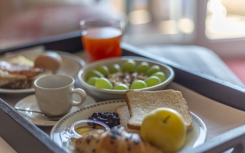 Close-up of a breakfast tray with grapes, granola, toast, apple, croissant, rolls, coffee, and orange juice.