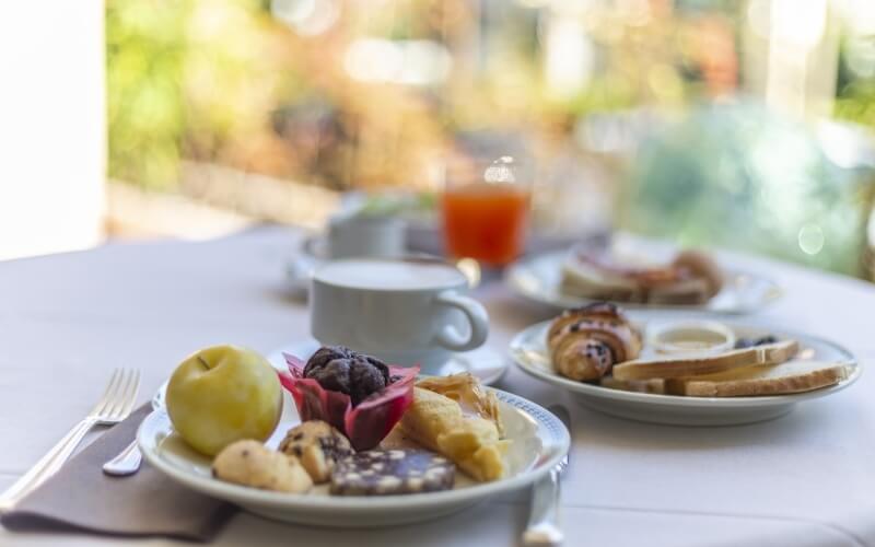 A breakfast table with a white tablecloth, pastries, fruit, coffee, orange juice, and cutlery arranged for a leisurely meal.