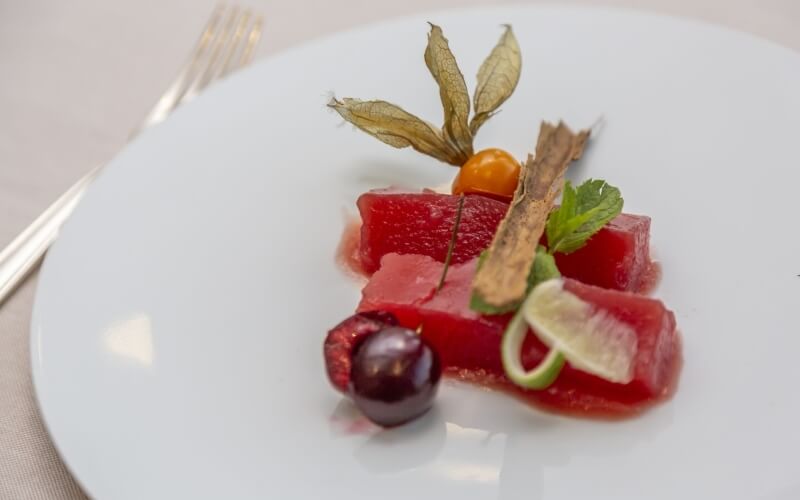 Close-up of a red gelatinous dessert on a white plate, garnished with fruits and herbs, with a blurred table setting background.