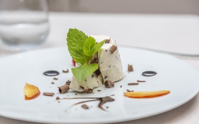 A creamy dessert with mint leaves, chocolate shavings, and fruit slices on a white plate, with a glass of water in the background.