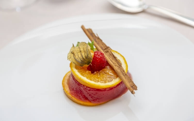 A circular dessert with red jelly and yellow-orange top, garnished with orange slice, raspberry, and green leaf on a white plate.