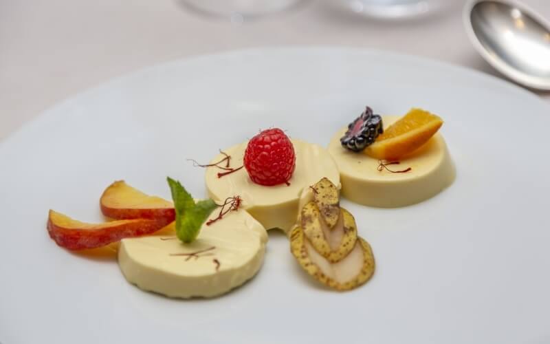 A round yellow dessert garnished with raspberry, orange, apple, mint, and saffron, on a white plate with a spoon nearby.