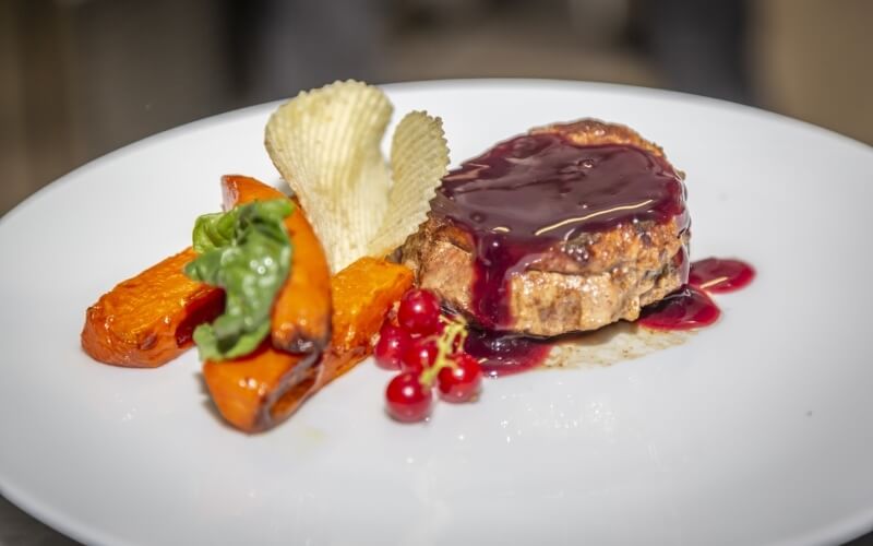 A plate with meat in red sauce, sweet potato slices, red currants, and waffle potato chips, showcasing vibrant colors and textures.