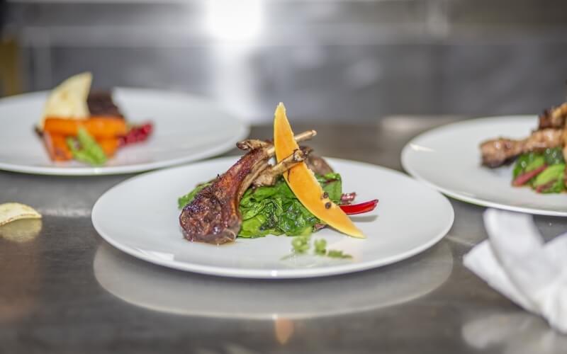 Three plates of food on a stainless steel surface; central plate features lamb chop, green vegetables, melon, and chili pepper.