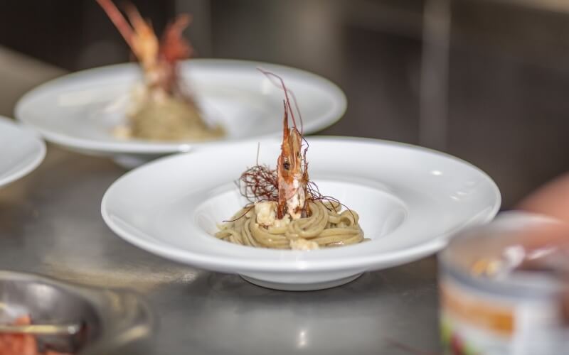 Three white bowls of shrimp-topped noodles on a stainless steel counter, suggesting a high-end dining experience.