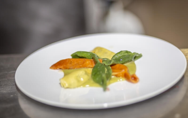 A white plate with colorful ravioli in green, orange, and yellow sauces, set on a blurred stainless steel kitchen counter.