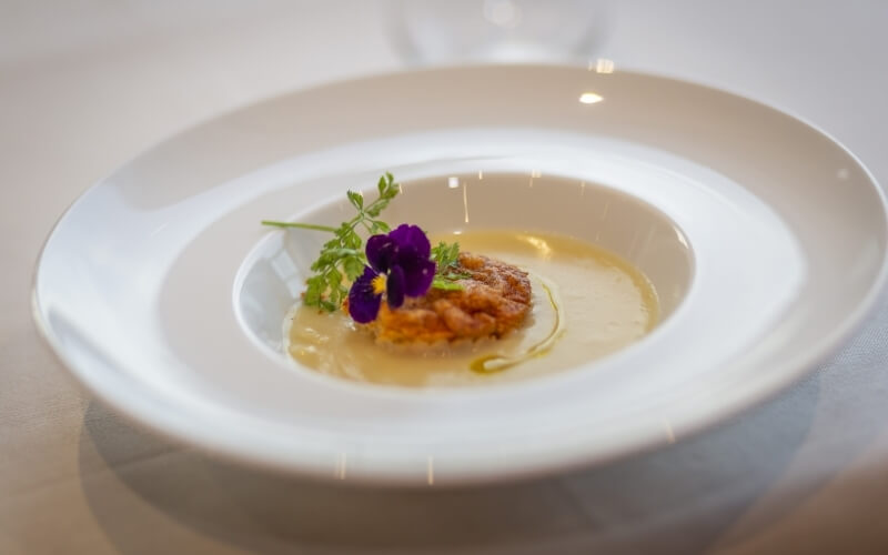 A shallow white bowl with pale yellow sauce, golden brown food, green herbs, and purple flowers, set on a blurred table.