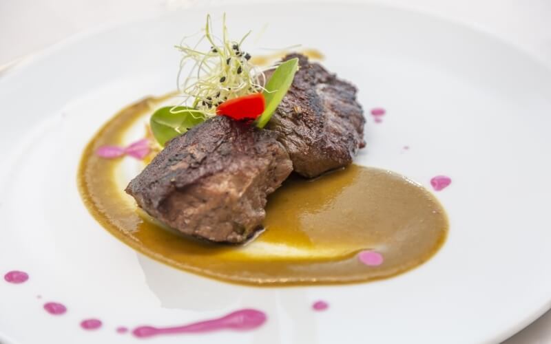 Elegant dish featuring two meat pieces on a dark and yellow sauce, garnished with red pepper, green leaf, and alfalfa sprouts.