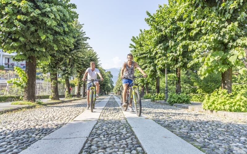 Two cyclists ride on a cobblestone path surrounded by greenery, with a clear blue sky and a white fence in the background.