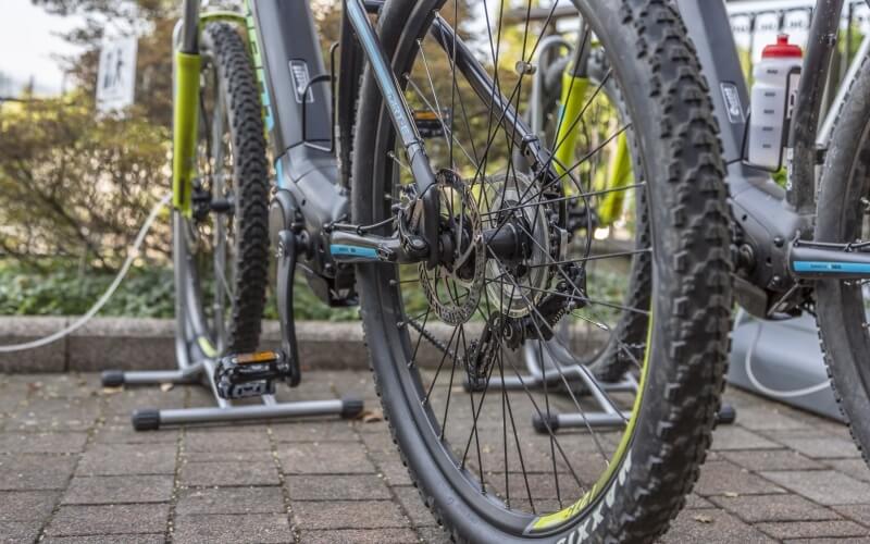 Close-up of a bicycle's front wheel on cobblestones, with another bike in the background and a water bottle attached.