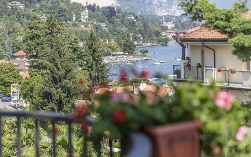 Scenic town view from a balcony with potted plants, overlooking water, boats, and buildings with white exteriors and terracotta roofs.