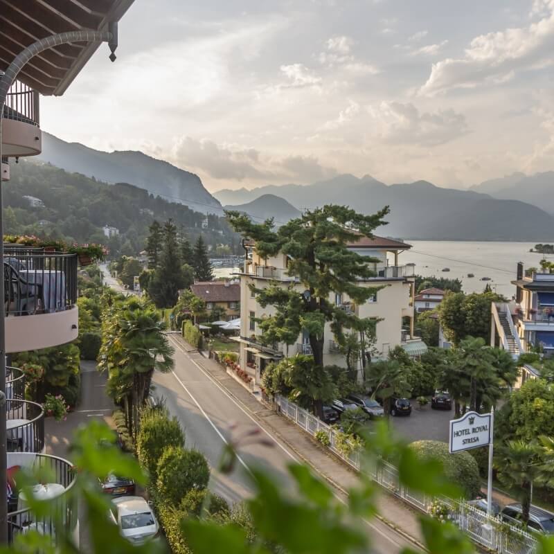 Serene hotel scene with "HOTEL ROYAL STRESA" sign, balconies, lush greenery, lake, and majestic mountains in the background.