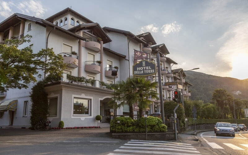 Multi-story hotel "HOTEL DELLA TORRE" with balconies, light facade, surrounded by trees and hills in a serene setting.