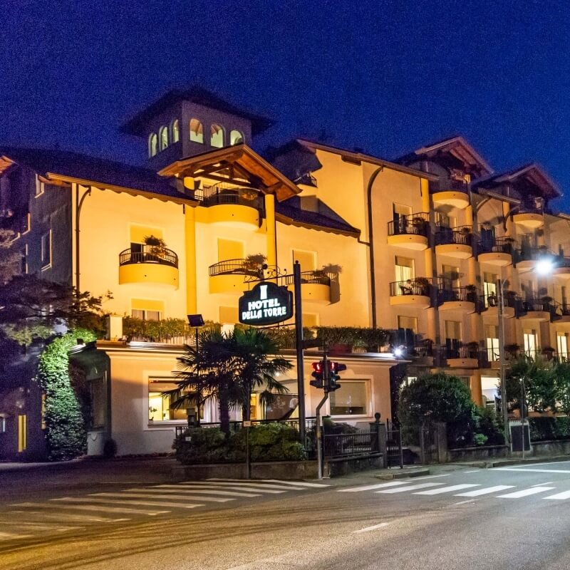 Multi-story hotel with white facade, balconies, "HOTEL DELLA TORRE" sign, landscaped area, and night sky.