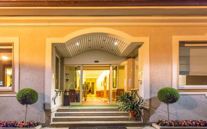 Welcoming beige building exterior with arched doorway, warm lighting, steps, windows, and potted plants.