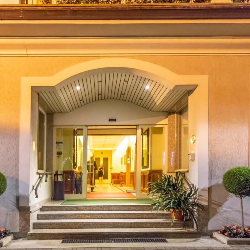 Ingresso di un hotel illuminato di notte, con lobby accogliente e persone visibili, circondato da piante e archi luminosi.