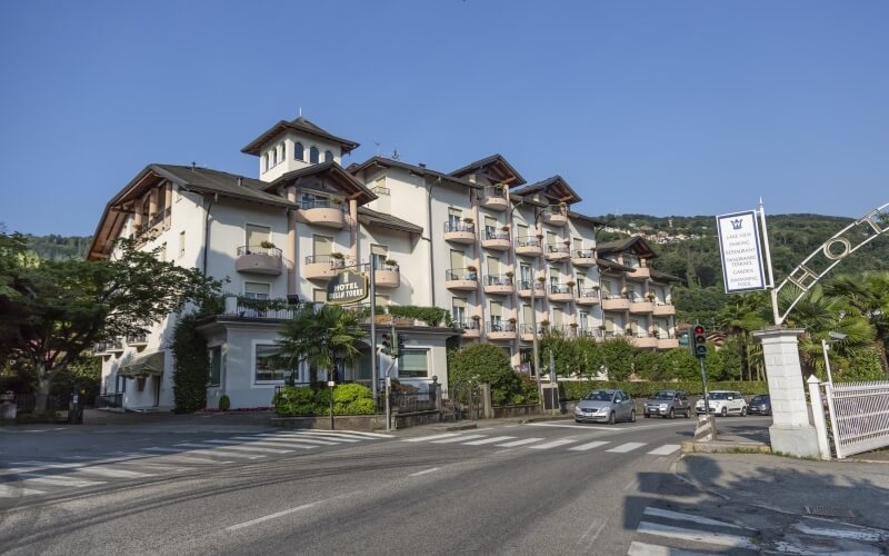 Multi-story white hotel with balconies, "HOTEL Sinstein" sign, parked cars, trees, and clear blue sky.