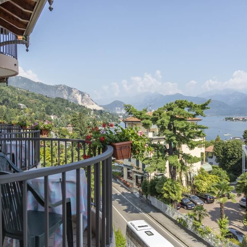 Balcony with table and chairs, potted flowers, scenic view of road, water, mountains, and blue sky on a sunny day.