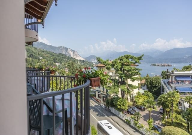 Balcony with black railing, potted plants, overlooking a town, lake, and mountains under a blue sky with clouds.