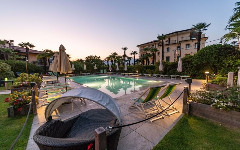 Serene poolside scene with a large pool, lounge chairs, a wicker chair, and a multi-story building amidst lush greenery.