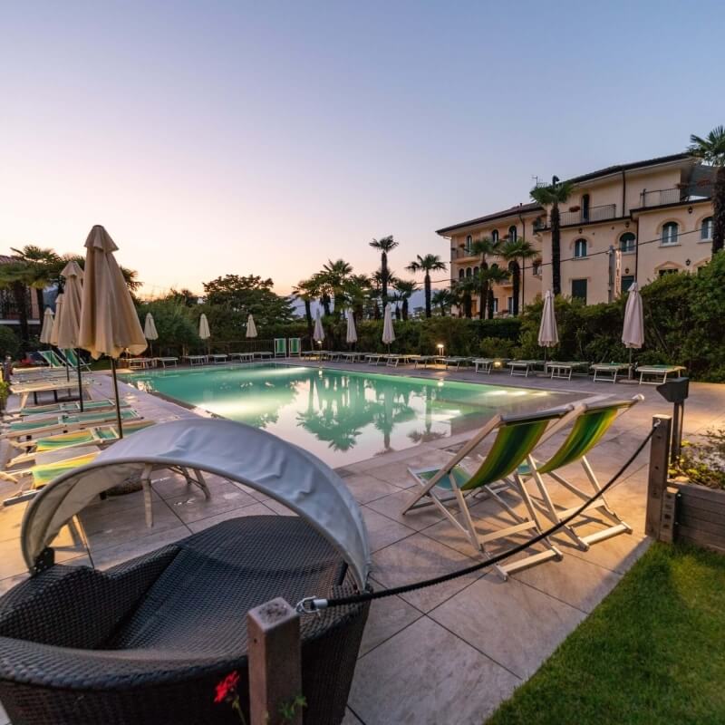 Serene poolside scene with a large rectangular pool, lounge chairs, umbrellas, palm trees, and a building in the background.