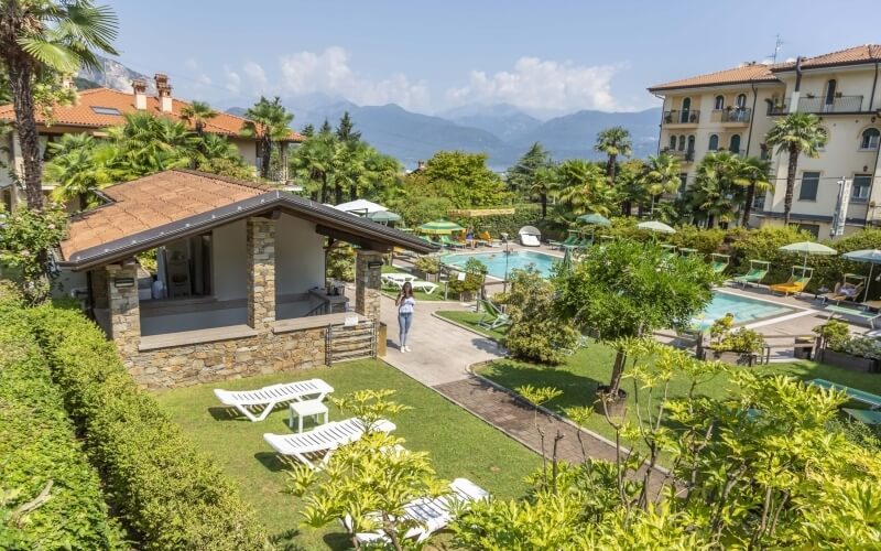 A woman walks towards the camera in a serene resort with a pool, lounge chairs, and mountains under a blue sky.