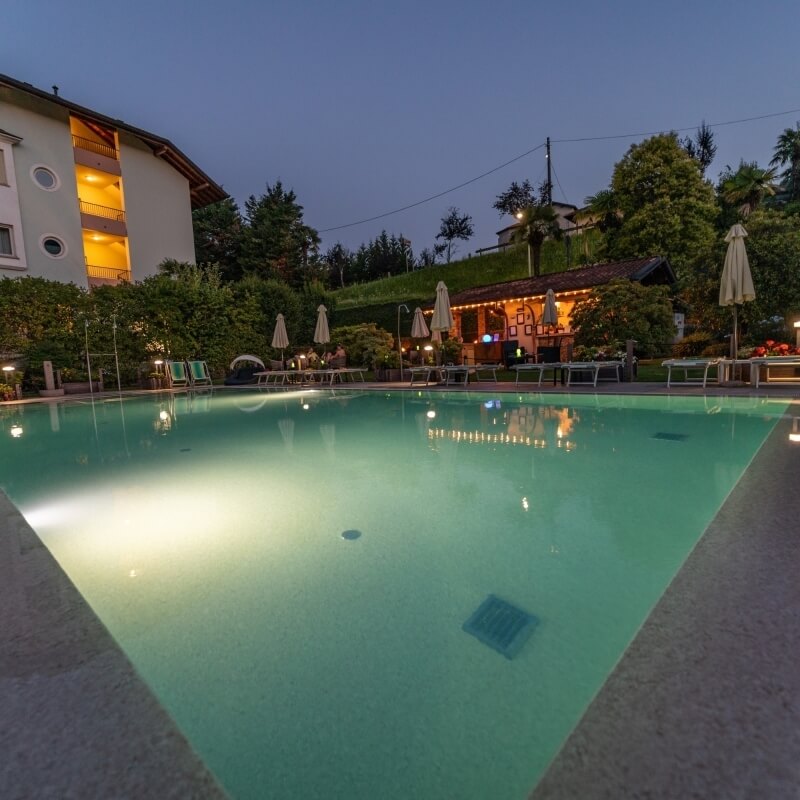 Serene dusk pool scene with light blue-green water, illuminated by underwater lights, surrounded by lounge chairs and trees.