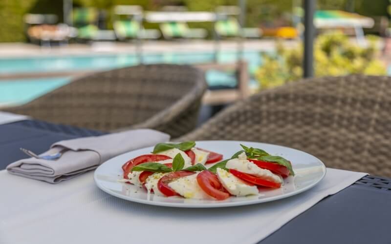 A plate of caprese salad on a table, with silverware, wicker chairs, and a swimming pool in a serene setting.