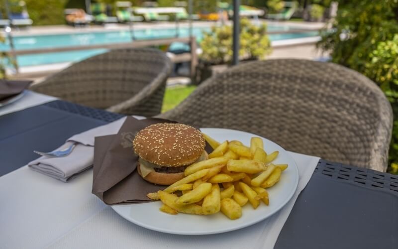 A white plate with a burger and fries on a tri-colored tablecloth, with a pool and lounge chairs in the background.