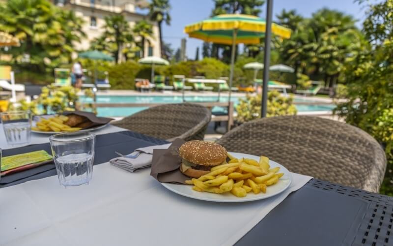 Table with a burger, fries, and water glasses in front of a pool with lounge chairs and umbrellas on a sunny day.