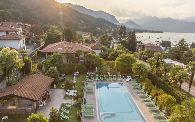 Serene poolside scene with lush greenery, terracotta-roofed houses, and a hot tub, set against mountains and a tranquil body of water.