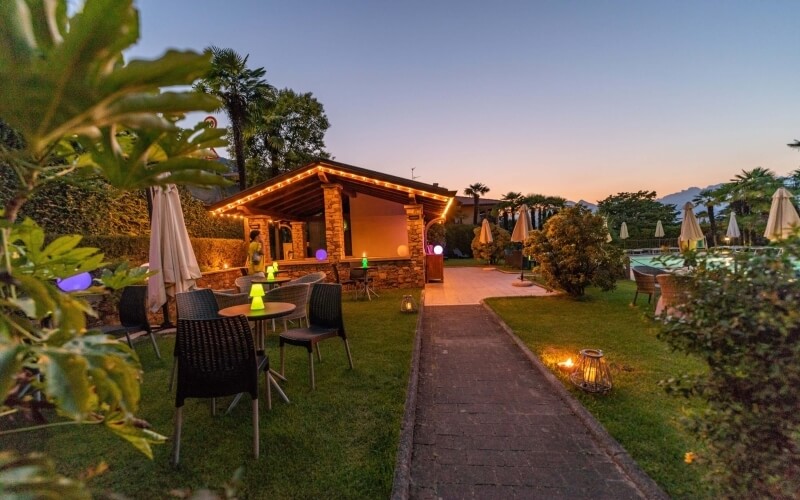 Serene outdoor setting with grassy area, tables with yellow lanterns, dark brown chairs, and a pathway to a lit building.