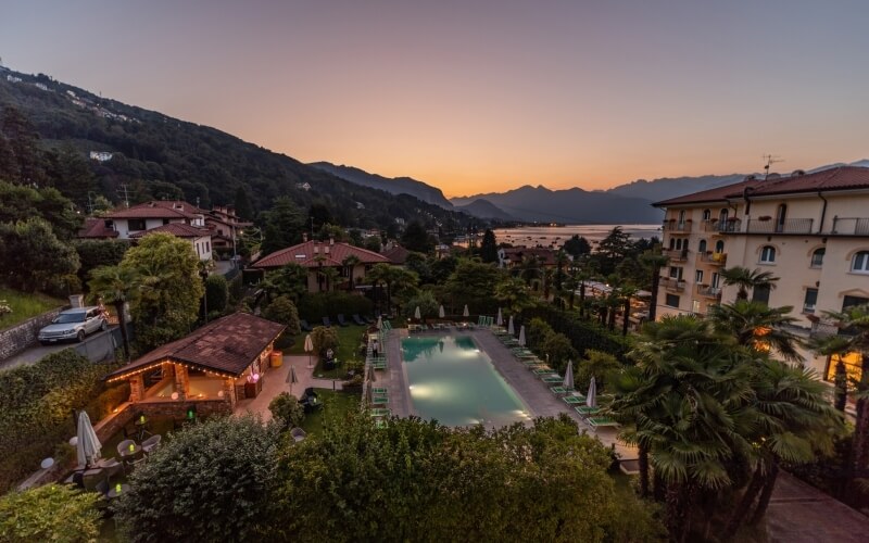 Serene resort scene with a rectangular pool, lounge chairs, and a warm sunset sky, surrounded by lush greenery and buildings.