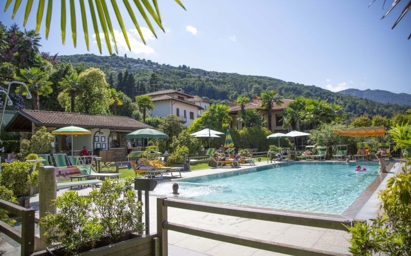 Serene pool scene with lush greenery, people lounging, and buildings in the background under a clear blue sky.