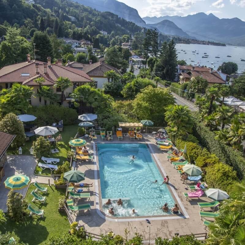 Aerial view of a serene pool area with a hot tub, surrounded by greenery, mountains, and a lake in the background.