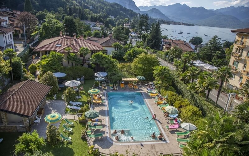 Outdoor swimming pool in a tropical setting, surrounded by lounge chairs, umbrellas, trees, and a distant lake with mountains.