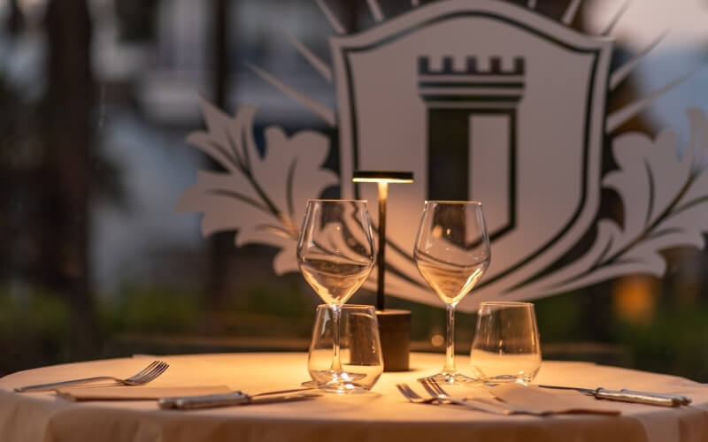 Dinner table for two with wine glasses and a shield logo in the background, suggesting an upscale dining atmosphere.