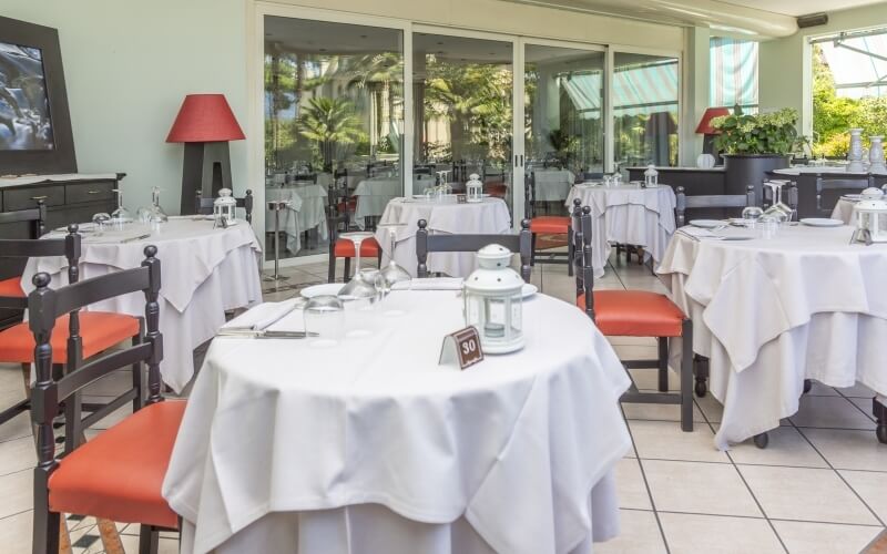 Restaurant dining area with white tablecloths, red chairs, large window wall, and a flat-screen TV on a sideboard.