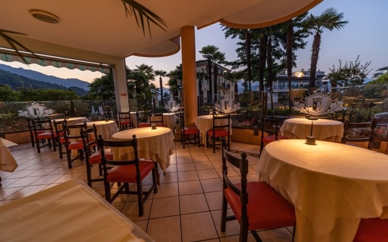 Interior of an elegant restaurant with round tables, red chairs, glass walls, and a view of a lush garden and mountains.