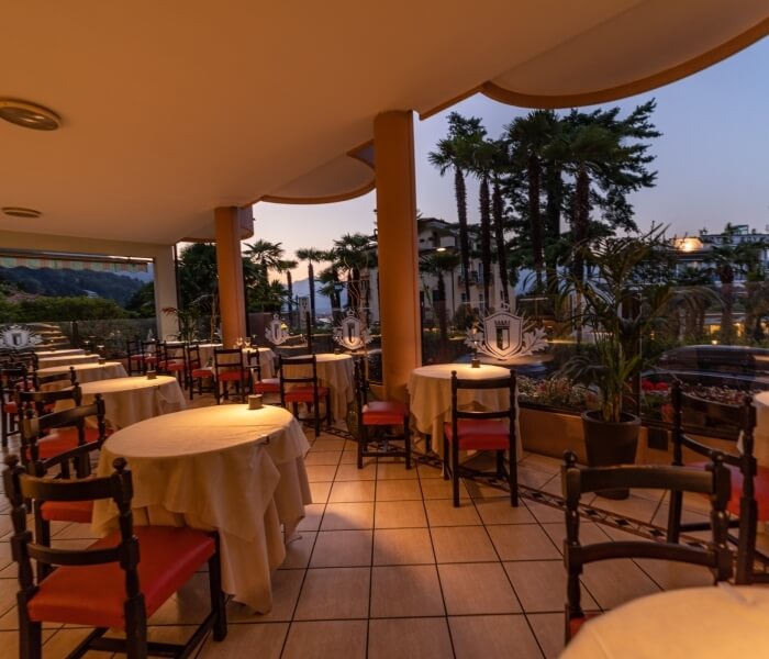 Outdoor seating area with tiled floor, white tablecloths, red chairs, palm trees, and a twilight sky with an orange glow.