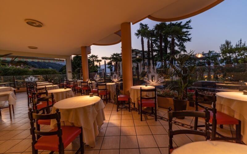 Open-air dining area with round tables, white tablecloths, red-cushioned chairs, and a serene landscape beyond a railing.