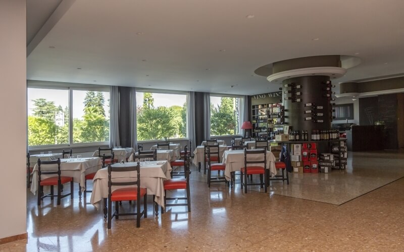 Elegant dining room with white walls, brown flooring, red-cushioned chairs, and a wine bar, surrounded by large windows.