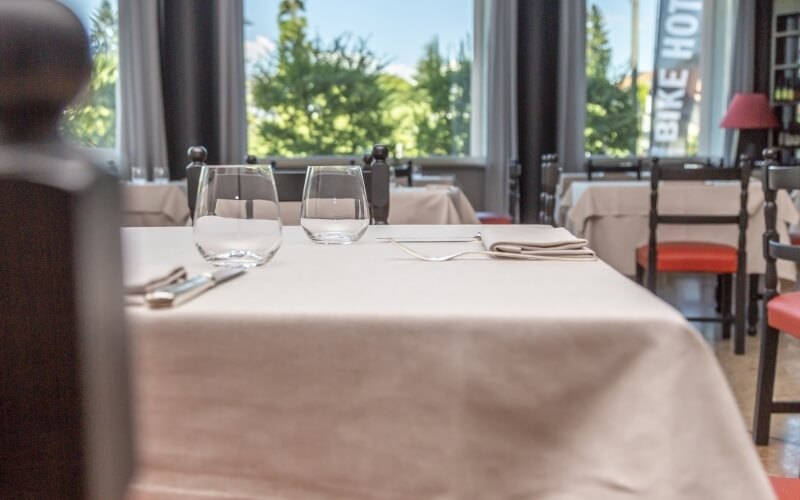 A dining room with tables set for service, featuring white tablecloths, glasses, napkins, and silverware, illuminated by natural light.