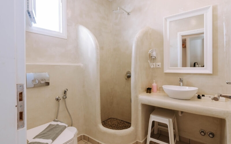 Bagno moderno con doccia, lavabo e WC, pareti beige, specchio bianco, luce naturale e sgabello sotto il lavabo.