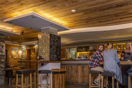 A cozy modern bar with wooden and stone decor, featuring patrons, a chalkboard menu, and a warm atmosphere.