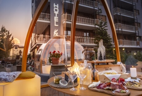 Outdoor dining setup with food and drinks in front of a large hotel, surrounded by greenery under a golden sky.