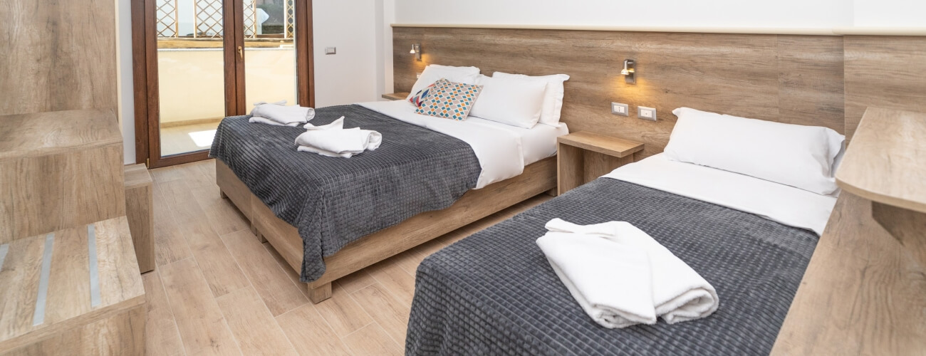 Cozy hotel room with two beds, light wood flooring, balcony door, and natural light from a window.