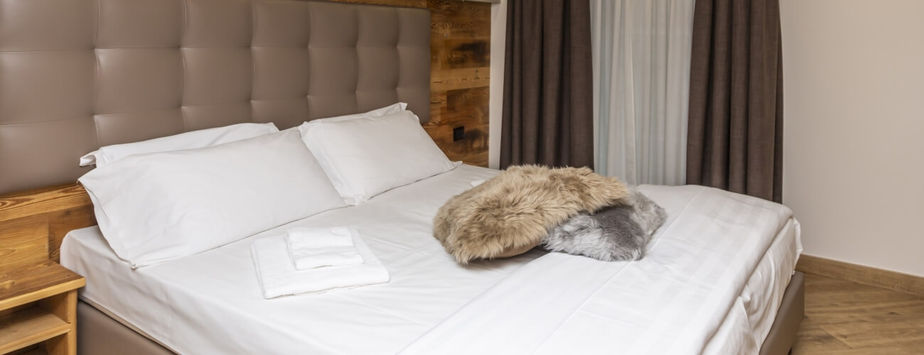 Serene bedroom with a tufted tan headboard, white comforter, nightstand, and elegant flowing curtains.