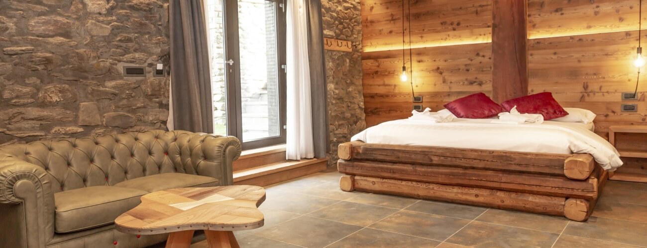 Camera rustica con parete in pietra, letto grande, divano in pelle tufted e tavolo in legno, illuminata da luce naturale.